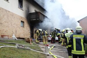 Einsatzkräfte vor dem brennenden Mehrfamilienhaus. Auf der rechten Seite steigt heller Rauch auf.