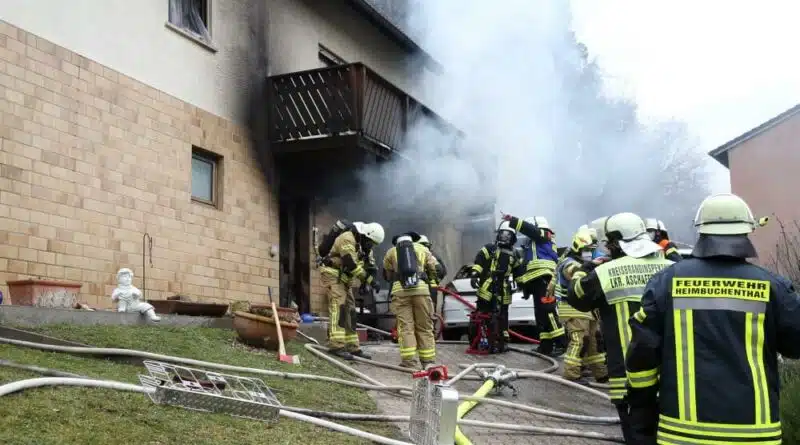 Einsatzkräfte vor dem brennenden Mehrfamilienhaus. Auf der rechten Seite steigt heller Rauch auf.