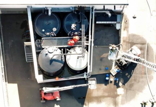 Draufsicht auf das Übungsszenario: Einsatzkräfte in roten Schutzanzügen suchen das Leck in den Rohrleitungen über den Tanks.